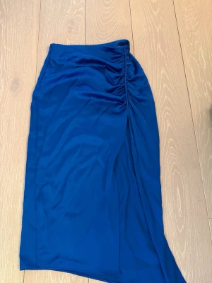 Blå långkjol med slits - Snygg blå långkjol med rynkdetalj och slits framtill. Kjolen har en smickrande passform och är gjord i ett mjukt, lätt material som ger ett elegant fall. Perfekt för dig som vill ha en stilren men ändå trendig look.