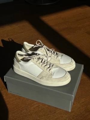 Vit/beiga Vagabond Teo i mocka och läder - Vita Vagabond Teo Sneakers med beiga partier. En stilren ”army trainer” inspirerad modell i skinn och mocka.   Endast använda två gånger och därav väldigt gott skick. Endast ett fåtal små märken på gummit. Original lådan ingår. Nypris för skorna var 1400 kr.  De är stora i storleken. Enligt avgivningar i skon och på kartongen är de storleken 41 men de passar egentligen storlek 43. Innermåttet är 27,5 cm. Jag själv har vanligtvis storlek 43 och de passar mig. 