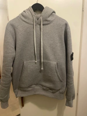Grå hoodie från Stone Island - Snygg grå hoodie från Stone Island med klassisk patch på ärmen. Hoodien har huva med dragsko och en stor magficka framtill. Tillverkad i mjukt bomullsmaterial och har ribbade muddar vid ärmslut och nederkant. Perfekt för en avslappnad streetwear-look.