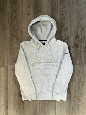 Ljusgrå hoodie från Superdry - Snygg ljusgrå hoodie från Superdry med broderad logga framtill och klassisk känguruficka. Hoodien har snörning i huvan och ribbade muddar vid ärmslut och nederkant. Perfekt för en avslappnad och trendig stil.