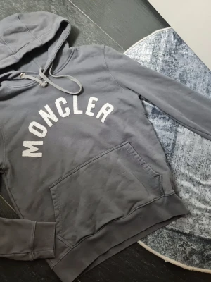 Grå Moncler hoodie med logga - Snygg grå hoodie från Moncler med stor vit logga i textur på bröstet. Klassisk modell med huva, dragsko och magficka. Perfekt för en chill och trendig look. Materialet känns mjukt och skönt mot huden.