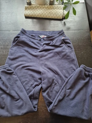 Blåa mjukisbyxor från Tommy Hilfiger - Mörkblå mjukisbyxor från Tommy Hilfiger med resår i midjan och vid bensluten. Byxorna har sidofickor och ett stort vitt Tommy-tryck längs ena benet. Tillverkade i mjukt bomullsmaterial för maximal komfort.