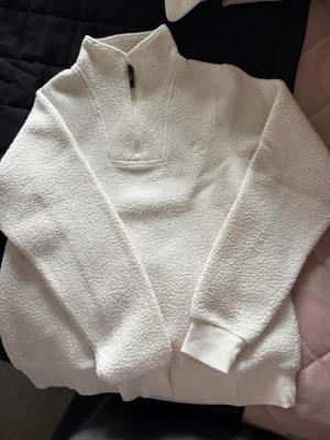 Vit teddytröja med halv dragkedja - Mysig vit teddytröja med halv dragkedja och hög krage. Tröjan har ribbade muddar vid ärmslut och nederkant, perfekt för lager-på-lager. Materialet är fluffigt och ger en cool streetkänsla.