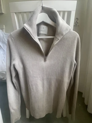 Beige ribbstickad tröja med halvzip - Snygg beige ribbstickad tröja från Jack and Jones med halvzip och hög krage. Tröjan har långa ärmar och en stilren, minimalistisk look som passar perfekt till många outfits. Materialet är mjukt och ger en skön känsla. Perfekt för dig som gillar klassisk men modern stil.
