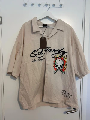 Beige Ed Hardy skjorta med tryck - Snygg beige skjorta från Ed Hardy med ikoniskt dödskalletryck med eldflammor och logga på bröstet. Köpt begagnad på Plick men har aldrig använts då den tyvärr är för stor för mig. Lappen sitter kvar!