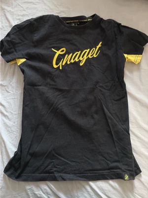 Svart Gnaget t-shirt med gul text - Storlek 146-152. Snygg svart t-shirt med gul text 'Gnaget' på bröstet och gula detaljer vid ärmsluten. Klassisk rund halsringning och normal passform. Perfekt för dig som vill visa ditt AIK-supporterhjärta. Tillverkad i mjuk bomull för skön känsla.