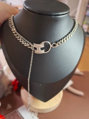 Silverfärgat halsband med H-berlock - Stilrent halsband i silverfärg med grov kedja och en glittrande H-formad berlock i mitten. Halsbandet har en extra kedjedetalj som hänger ner och ger en trendig look. Perfekt accessoar för att lyfta din outfit.