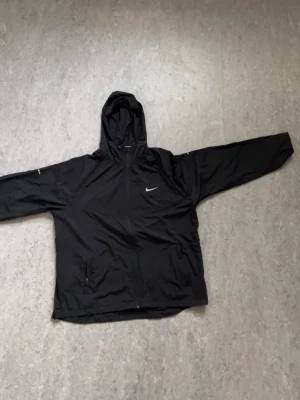 Svart Nike vindjacka med huva - Svart vindjacka från Nike med huva och dragkedja. Jackan har reflekterande detaljer på bröstet och ärmarna samt två fickor framtill. Perfekt för blåsiga dagar och sportiga outfits.