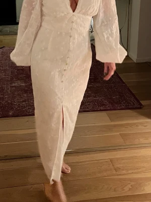 Vit långklänning med broderade blommor - Superfin vit långklänning med djup v-ringning, långa puffärmar och knappar framtill. Klänningen har broderade blommor och blad över hela tyget och är gjord i ett lätt, luftigt material. Perfekt för studenten och sommarens alla tillfällen!