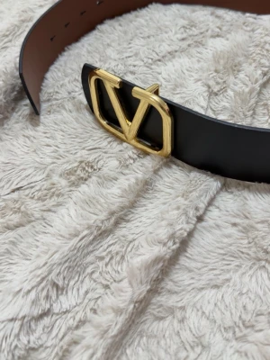 Svart skärp med guldfärgat V-spänne - Stilrent svart skärp från Valentino med ett stort guldfärgat V-logga som spänne. Bältet är gjort i slätt skinn och har en klassisk, tidlös design som lyfter vilken outfit som helst.
