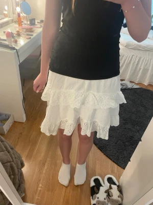 Vit volangkjol med broderade detaljer - Superfin vit kortkjol med flera volanger och broderade spetsdetaljer. Kjolen har en luftig och söt design som passar perfekt till varmare dagar. Tillverkad i bomull för en skön och lätt känsla.