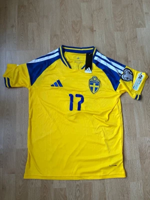 Sveriges Hemma matchtröja Adidas 2026 - Sveriges Hemma matchtröja från Adidas med nummer 17 på bröstet och officiellt landslagsmärke med Gyökeres på ryggen. Tröjan har blå detaljer på axlar och ärmar, samt broderad Adidas-logga. Tillverkad i lätt och ventilerande material, perfekt för fotboll. Helt oanvänd med lappar kvar, skriv vid frågor eller funderingar.
