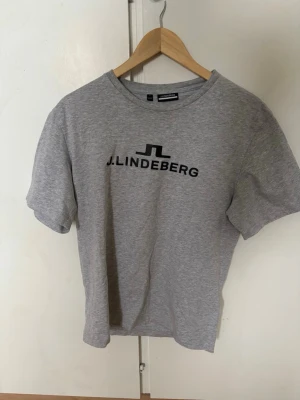 Grå t-shirt från J.Lindeberg - Snygg grå t-shirt från J.Lindeberg med svart logotyptryck på bröstet. Klassisk rund halsringning och korta ärmar. Perfekt för en stilren och avslappnad look.