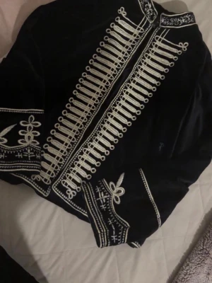 Zara limited edition napoleon jacka - Unik svart sammetsjacka från grf med detaljerad vit brodyr i militärstil. Jackan har ståkrage, dekorativa knappar och mönster längs ärmar och framstycke. Perfekt för dig som vill sticka ut med en cool och retroinspirerad look.