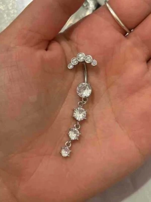 Silvrigt navelpiercingsmycke med stenar - Säljer ett silvrigt navelpiercingsmycke med flera gnistrande, runda stenar i rad. Smycket har en böjd stav och hängande design med fyra mindre stenar under en större. Perfekt för dig som vill ha något glittrigt och stilrent.