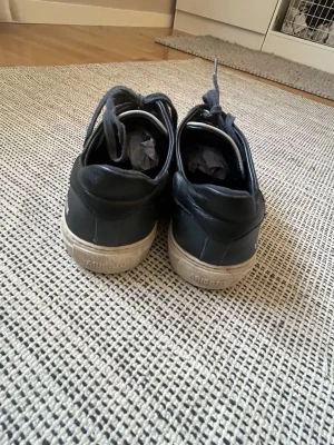 Svarta sneakers från Axel Arigato - Snygga svarta sneakers från Axel Arigato med vita sulor och mörkblå detaljer. Skorna har låga skaft, svarta skosnören och är tillverkade i läder och mocka. Perfekta för dig som gillar stilrena och klassiska sneakers.