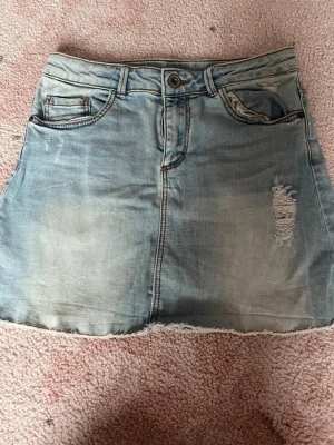 Ljusblå jeanskjol med slitningar - Snygg ljusblå jeanskjol från Zara med slitna detaljer och rå kant nertill. Kjolen har klassisk femficksdesign, knapp och dragkedja framtill samt en liten slits bak. Perfekt för en avslappnad och trendig look.