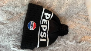 Svart pepsi mössa - Säljer en svart stickad mössa med vit text där det står PEPSI och en klassisk Pepsi-logga framtill. 