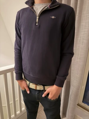Mörkblå halfzip tröja från Gant - Mörkblå sweatshirt från Gant med halv dragkedja och ståkrage. Tröjan har diskret Gant-logga broderad på bröstet och ribbade muddar vid ärmslut och nederkant. Perfekt för en avslappnad men snygg stil.