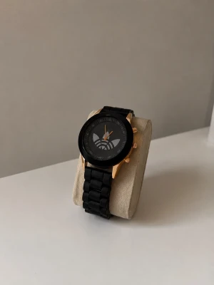 Svart klocka från Adidas med gulddetaljer - Stilren svart klocka från Adidas med bred länk och tydlig logga på urtavlan. Klockan har guldfärgade detaljer på boetten och knapparna, vilket ger en snygg kontrast till det svarta bandet. Perfekt accessoar för en sportig och modern look.