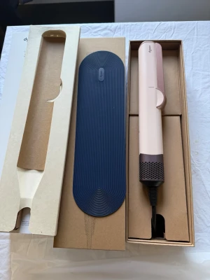 Dyson Airstrait hårstyler rosa - Dyson Airstrait hårstyler i ljusrosa och grått. Kommer med originalförpackning, värmetålig matta och laddare. Perfekt för att styla och torka håret snabbt med hjälp av luft istället för värmeplattor. Snygg och modern design i plast och metall.