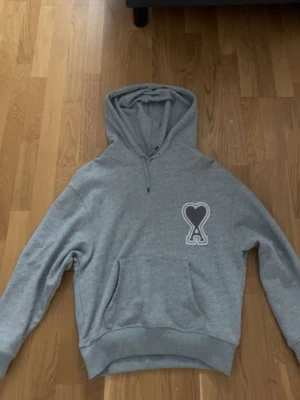 Grå hoodie från Ami Paris med hjärtdetalj - Säljer en grå hoodie från Ami Paris med deras ikoniska hjärta och A-logga på bröstet. Hoodien har huva med dragsko och en stor magficka. Tillverkad i mjukt bomullsmaterial och har ribbade muddar vid ärmslut och nederkant. Perfekt för en chill och stilren look. Den passar också med M