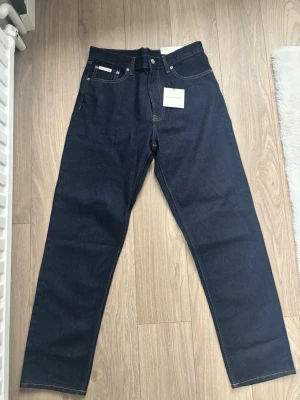 Mörkblå 90s Taper jeans Calvin Klein - Snygga mörkblå jeans från Calvin Klein Jeans i modellen 90s Taper. Klassisk femficksdesign, raka ben och orange kontrastsömmar. Perfekta för dig som gillar en retroinspirerad look med modern passform.