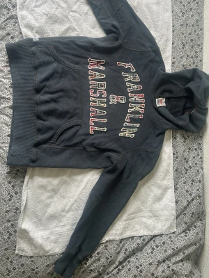Mörkblå Franklin & Marshall hoodie - Snygg mörkblå hoodie från Franklin & Marshall med stor logga i vitt och rött på bröstet. Klassisk känguruficka framtill och ribbade muddar vid ärmar och midja. Tillverkad i mjukt bomullsmaterial och har en skön huva.