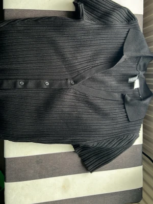 Svart ribbad kortärmad topp med krage - Svart ribbad topp med korta ärmar, v-ringning och klassisk krage. Toppen har knappar framtill och är i en mjuk, stretchig kvalitet. Perfekt att styla med jeans eller kjol. Är åtsittande. 