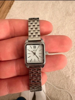 Regal silverfärgad fyrkantig klocka - Stilren klocka från Regal med fyrkantig urtavla i vitt och silverfärgat armband i metall. Klassisk design med romerska siffror och justerbar länk, extra länkar medföljer. Perfekt accessoar för dig som gillar enkel och tidlös stil.