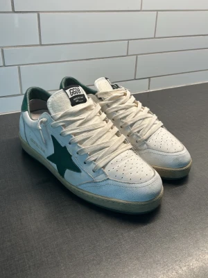 Vita Golden Goose Ball Star sneakers - Snygga vita Golden Goose Ball Star sneakers med gröna detaljer och stjärna på sidan. Skorna har gröna sulor undertill och är tillverkade i läder med klassisk låg siluett. Perfekta för dig som gillar streetstyle och unika detaljer.
