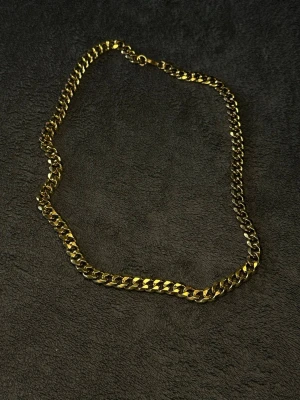 Guld Cubansk Kedja 55cm 9mm i Rostfritt Stål  - 55cm Långt 9mm tjockt halsband i guld färg. Stilrent halsband med breda, platta länkar i guldfärgat stål. Kedjan har en klassisk karbinhake och en glansig finish som ger en modern vibe. Perfekt för att lyfta din outfit med en edgy touch.