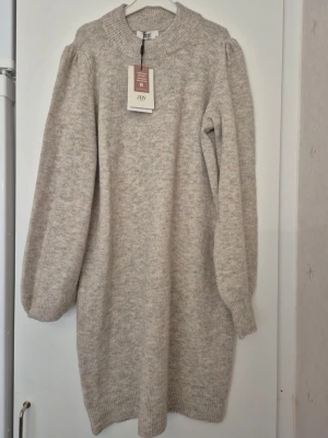 Klänning - Mysig mjuk beige/grå stickad klänning från JDY med rund halsringning och långa ärmar. Klänningen har en rak passform och ribbade muddar vid ärmslut och nederkant. Perfekt för kyligare dagar och enkel att styla med boots eller sneakers. Nypris: 300kr.