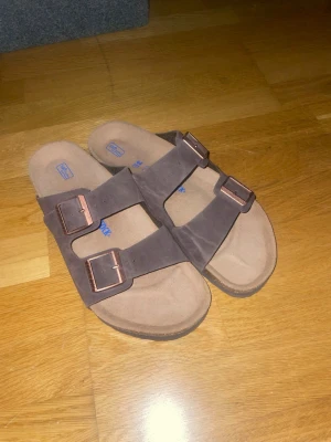 Bruna Birkenstock sandaler med spännen - Säljer ett par klassiska bruna Birkenstock sandaler med två breda remmar och snygga kopparfärgade spännen. Ovandelen är i mockaliknande material och sulan är mjuk och bekväm. Perfekta för chill dagar och enkel stil.