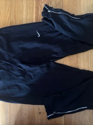 Svarta Nike byxor - Svarta byxor från Nike. Byxorna har dragkedjor vid bensluten och en liten vit Nike-logga på låret. Perfekta för chill eller träning, med snörning i midjan för bästa passform.