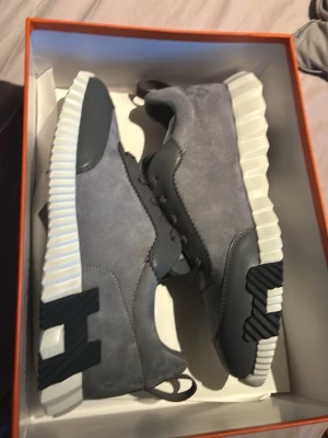 Grå sneakers med vit sula - Snygga sneakers från Hogan i grå mocka och läder med vita, räfflade sulor och svarta detaljer. Skorna har svarta skosnören och en modern, chunky design med tydlig H-logga på sidan. Perfekta för dig som gillar stilrena och trendiga sneakers.