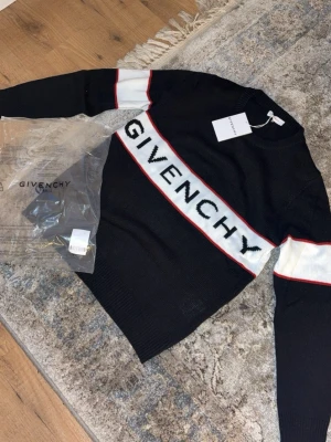 Svart stickad tröja från Givenchy - Snygg svart stickad tröja från Givenchy med vit panel och röd kant längs bröstet och ärmarna. Stor Givenchy-logga framtill. Rund halsringning och långa ärmar. Perfekt för dig som gillar exklusiva streetwear-plagg.