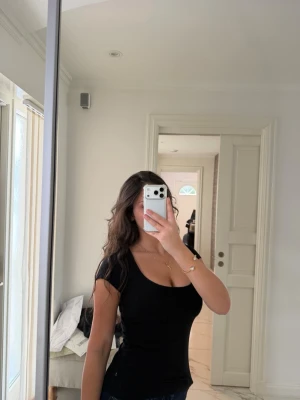 Svart tight topp med kort ärm - Snygg svart topp med tight passform och korta ärmar. Djup rundad urringning framtill som ger en modern look. Perfekt att matcha med jeans eller kjol för en stilren outfit.