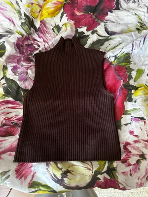 Brun ribbad ärmlös polotröja - Säljer en brun ribbstickad ärmlös polotröja med hög krage. Tröjan har en rak passform och är perfekt för lager på lager. Materialet känns mjukt och stretchigt, vilket gör den bekväm att bära.