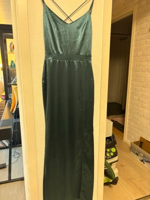 Mörkgrön balklänning. med slits -  I mjukt satinliknande material. Med fin spetsdesign på ryggen. Kontakta för fler bilder. 