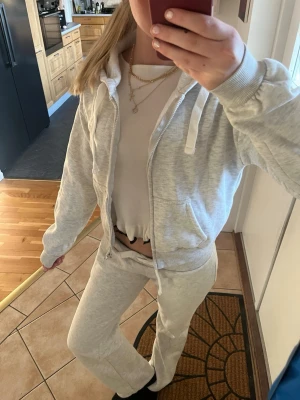 Ljusgrå mysig loungewear-set. Byxor och kofta - Loungewear-set i ljusgrå färg med zip-hoodie och matchande mjukisbyxor. Hoodien har dragkedja, fickor och vita snören, medan byxorna har resår och snörning i midjan. Perfekt för chill dagar hemma. Materialet är mjukt och bekvämt, troligen bomullsblandning.