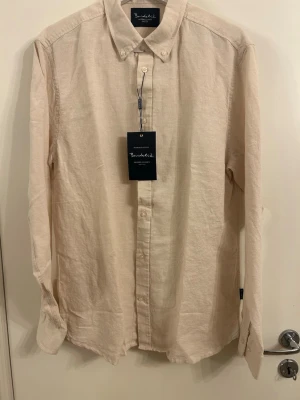 Beige linneskjorta från Bondelid  - Stilren beige skjorta i mjukt linne. Skjortan har klassisk krage, knappar framtill och långa ärmar med knapp vid manschetten. Perfekt för en avslappnad men snygg look. Aldrig använd prislapp kvar 
