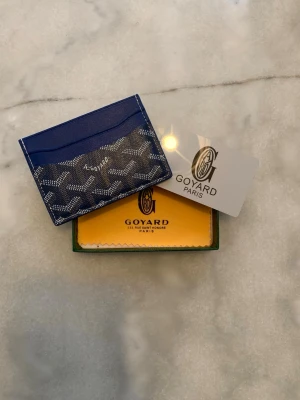 Blå korthållare från Goy4ard - Snygg blå korthållare från Goy4ard med det klassiska vita och gråa Goy4ard-mönstret. Tillverkad i canvas och läder, har flera fack för kort och är perfekt för dig som vill ha en stilren och exklusiv accessoar. Levereras med originalask och tygpåse.