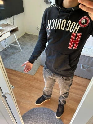  HOODRICH hoodie med tryck - Svart hoodie från HOODRICH med stor vit logga och röd H på bröstet. Har huva med dragsko, känguruficka och flera patches och tryckdetaljer. Perfekt för en streetwear-look och riktigt skön passform.