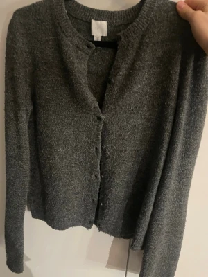 Mörkgrå kofta från H&M - Mysig mörkgrå kofta från H&M med rund halsringning och knappar framtill. Koftan har lång ärm och är stickad i ett mjukt material.