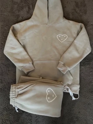 Beige mjukisset med hjärtdetalj - Mysigt beige loungewear-set med hoodie och matchande byxor. Båda plaggen har en vit broderad hjärtdetalj med smiley på bröstet och benet. Hoodien har känguruficka och byxorna har dragsko i midjan. Perfekt för chill dagar hemma.