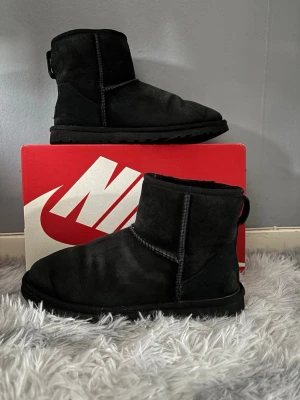 Svarta UGG tofflor i mocka 41 - Mysiga svarta tofflor från UGG i mjuk mocka med kort skaft och grov sula. Klassisk design med synliga sömmar och rundad tå. Perfekta för att hålla fötterna varma under kalla dagar. Använda men i bra skick