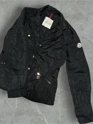 Vintage Svart vårjacka från Moncler - Snygg svart pufferjacka från Moncler med guldfärgade knappar och dragkedjor. Jackan har lång ärm, ståkrage och Moncler-logga på ärmen. Perfekt för kyliga dagar och ger en stilren look. Storlek 1 vilket motsvarar S. För dam