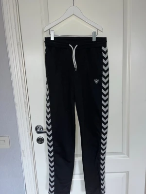 Svarta mjukisbyxor från Hummel - Säljer ett par helt nya svarta mjukisbyxor från Hummel med vita sicksackränder längs sidorna och dragsko i midjan. 