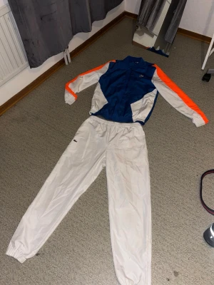  träningsdress från lacoste - Säljer en retro träningsdress från lacoste med blå och vita partier samt orange detaljer på ärmarna. Jackan har dragkedja och byxorna har resår i midjan och benslut. Perfekt för dig som gillar vintage sportstil.